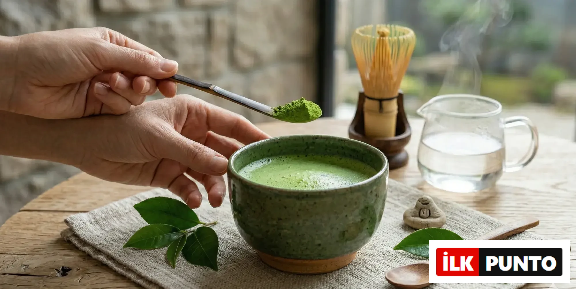 Seramik bir kasede hazırlanan matcha çayı; kaşıkla eklenen yeşil matcha tozu, yanında bambu çırpıcı, cam sürahide sıcak su ve doğal ahşap zemin üzerinde sakin bir ritüel atmosferi.