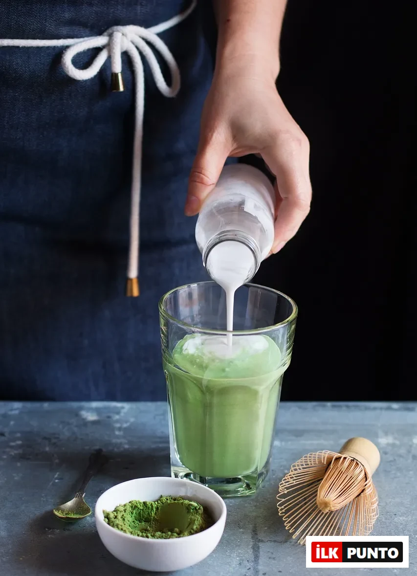 Bardakta hazırlanan matcha latteye süt eklenirken; yanında matcha tozu, küçük kaşık ve bambu çırpıcıyla sade ve modern bir hazırlık anı.