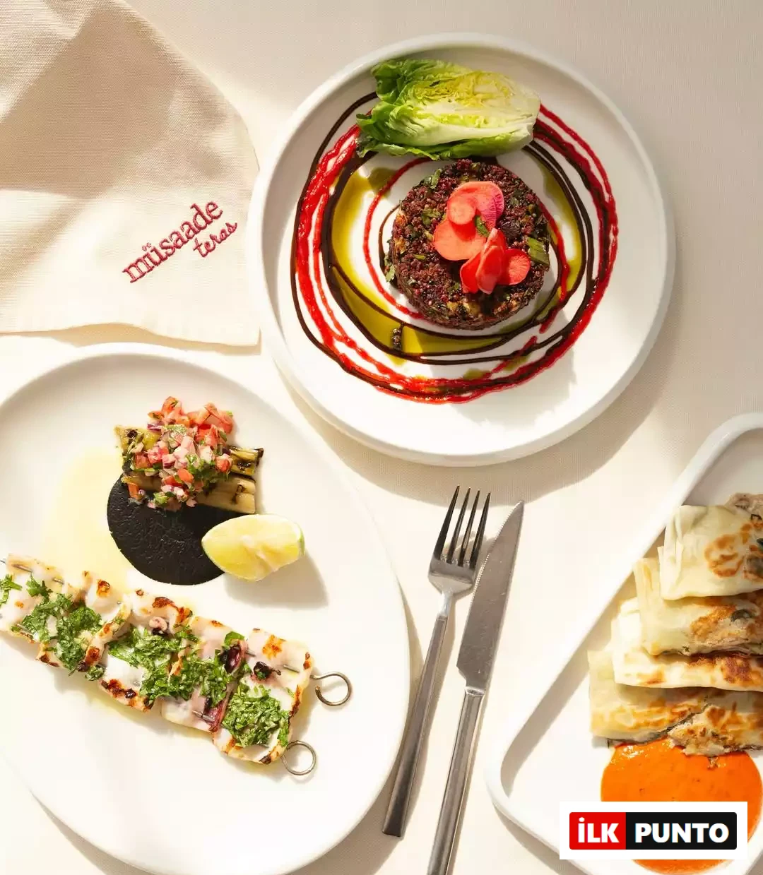 Şefin favori meyhanesi Müsaade Teras'ın modern meze masası. Parlak gün ışığında beyaz tabaklarda kırmızı soslu kinoa salatası, siyah soslu deniz ürünü şişi, börekler ve işlemeli mekan peçetesi zarif bir lezzet deneyimini