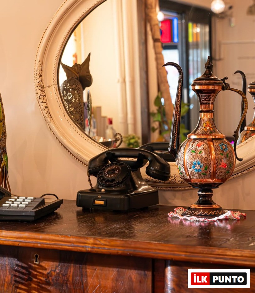 Koyu renk ahşap bir komodinin üzerinde duran eski tip siyah çevirmeli telefon ve el işi oymalı bakır ibrik, arka plandaki oval aynanın yansımalarıyla birleşerek Salaş Meyhane'nin Ankara'daki samimi ve geleneksel meyhane kültürünü gözler önüne seriyor.