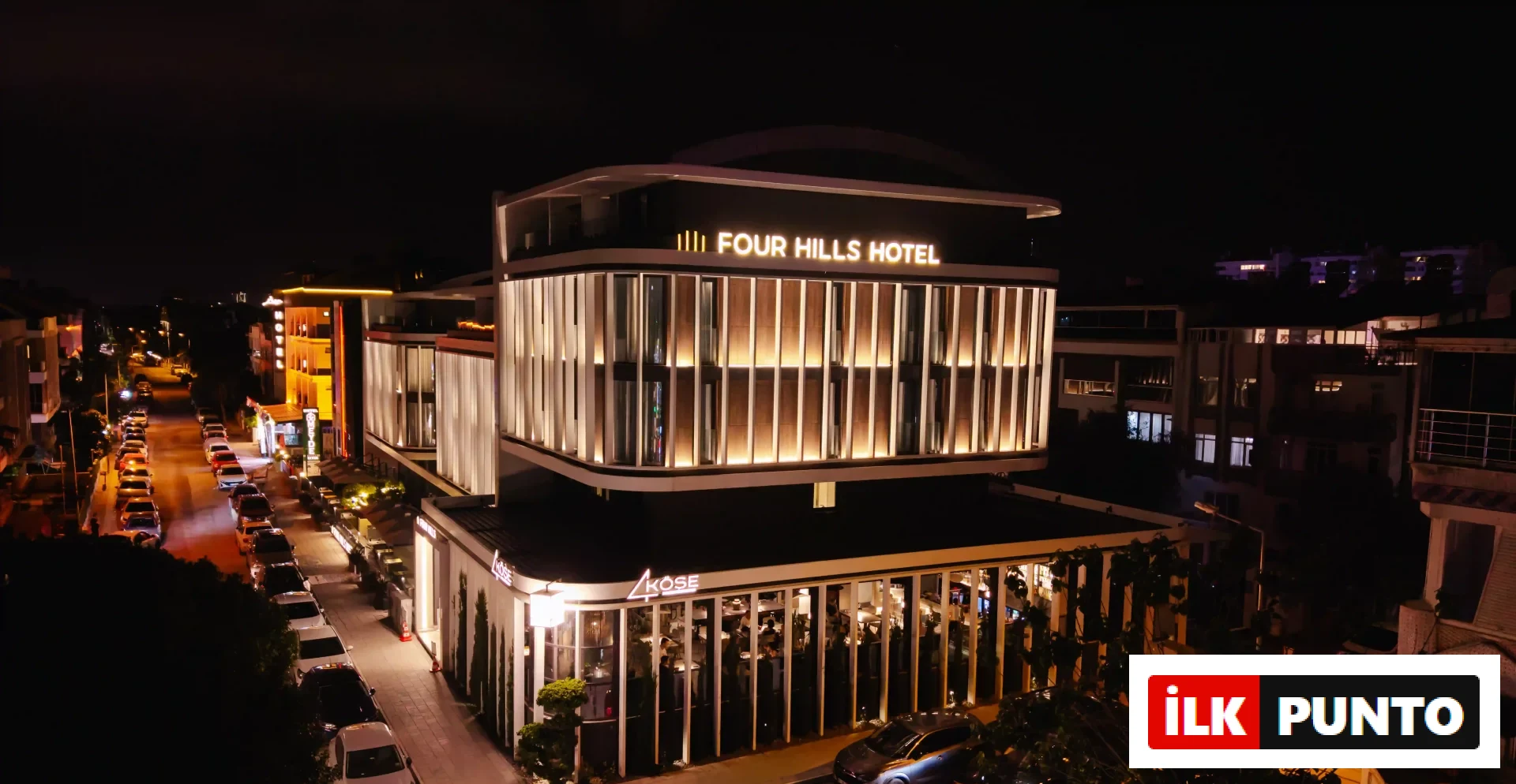 Antalya'da, gecenin karanlığında modern aydınlatmasıyla parlayan, üst katlarında Four Hills Hotel'i barındıran ve zemin katında misafirlerini ağırlayan şık KÖŞE restoranının geniş açılı bir görüntüsü; cadde boyunca park etmiş araçlar ve uzakta seçilen şehir ışıklarıyla tamamlanıyor.