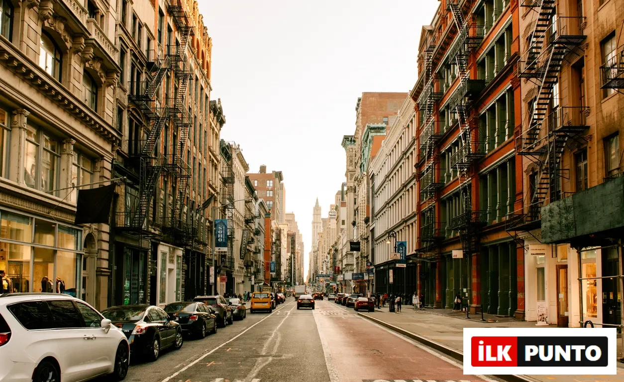 New York şehrinin hareketli bir caddesi, iki yanında dış yangın merdivenleriyle bezeli yüksek binaları, yol kenarında park etmiş çok sayıda arabası ve kaldırımlarda yürüyen insanlarıyla tipik bir kentsel manzarayı gözler önüne seriyor.