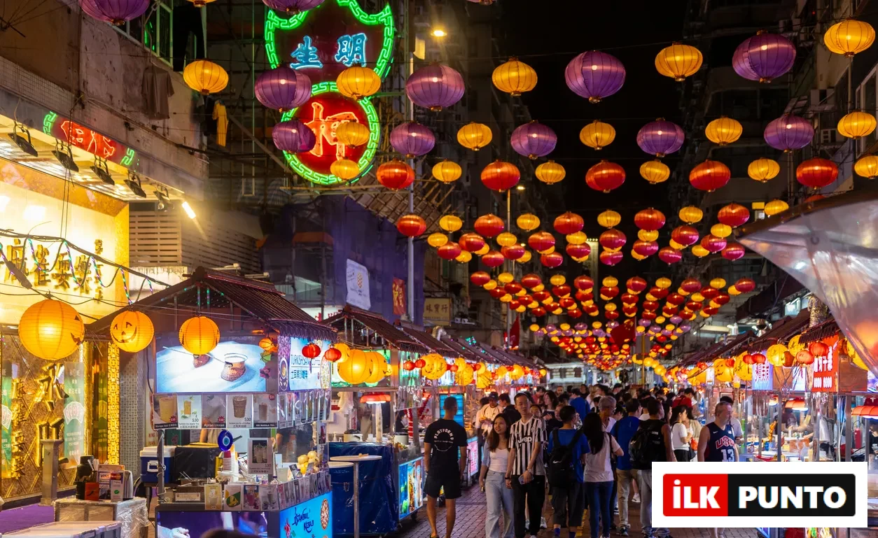 Hong Kong'da, gece vakti sokakları aydınlatan sarı, mor, kırmızı ve turuncu rengarenk fenerlerle süslenmiş, çeşitli yiyecek ve ürünler sunan tezgahlar arasında dolaşan kalabalıklarla dolu canlı bir gece pazarı manzarası.
