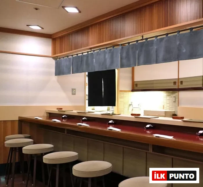 Sukiyabashi Jiro gibi ünlü bir suşi restoranının, zarif ahşap tezgahı boyunca düzenlenmiş altı minderli taburesi ve her biri için özenle yerleştirilmiş servis setleriyle geleneksel Japon zanaatkarlığını yansıtan iç mekanı.
