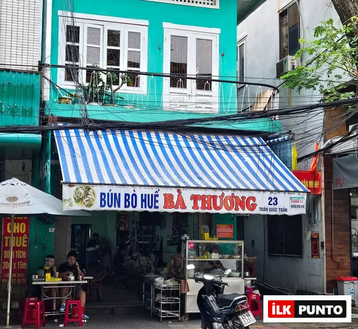 Bún Bò Huế Bà Thương adlı Vietnam restoranının turkuaz renkli binasının önünde, mavi beyaz çizgili tentenin altında masalarda oturup yemek yiyen insanları, yiyecek standını ve parkedilmiş bir motosikleti çevreleyen yoğun elektrik kablolarıyla dolu hareketli bir sokak görünümü.