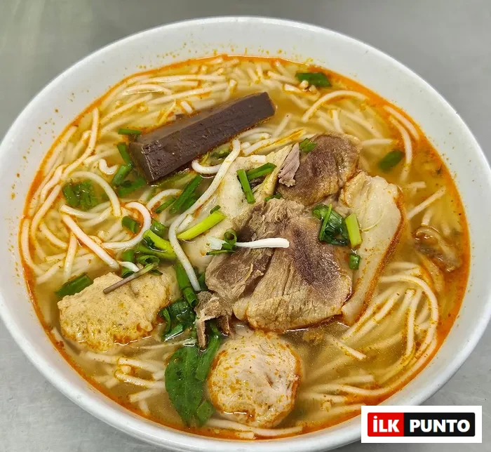 Yakın planda, Bun Bo Hue çorbası; beyaz pirinç eriştesi, dana ve domuz eti parçaları, köfteler, koyu renkli jöleleşmiş kan keki ve taze yeşil soğanlarla süslenmiş baharatlı kırmızımsı et suyunda iştah açıcı bir şekilde duruyor.