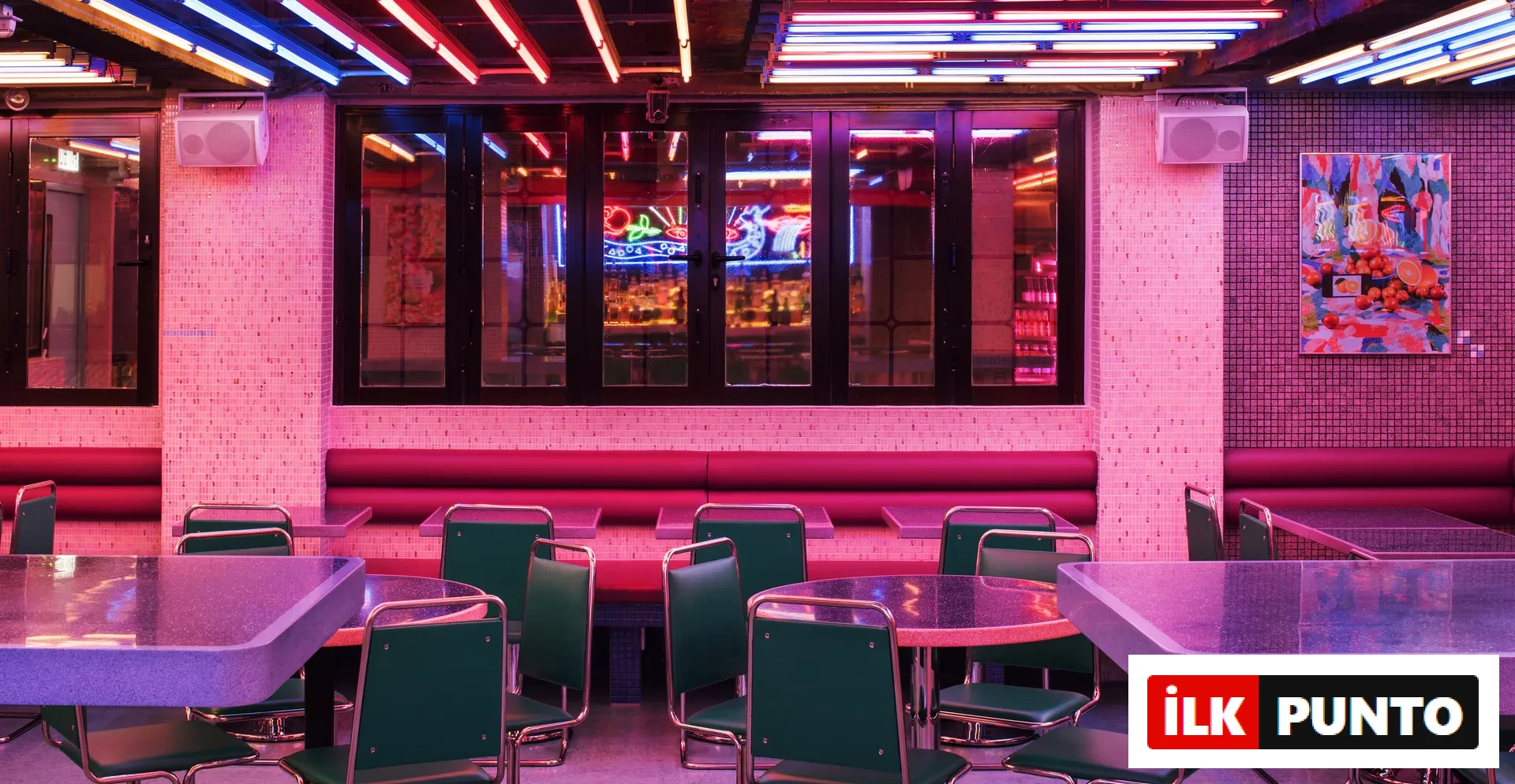 Parlak pembe ve mavi neon ışıkların hakim olduğu, pembe küçük kare fayanslarla kaplı duvarlar, bordo banklar ve yeşil krom sandalyelerle döşenmiş, yuvarlak ve dikdörtgen koyu renkli parıltılı masaların çevrelediği, enerjik bir restoran veya bar iç mekanı.