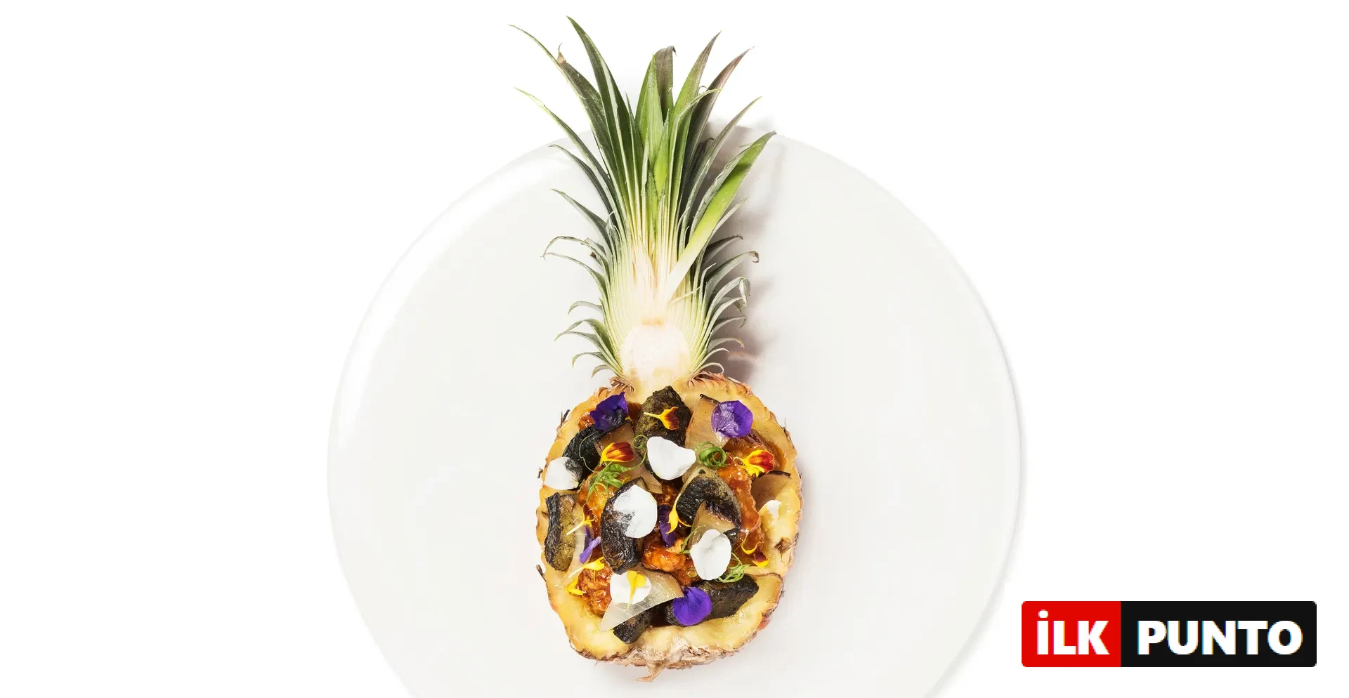 Beyaz bir tabakta, içi çeşitli malzemelerle doldurulmuş, zarifçe hazırlanmış yarım bir ananas yemeği; içerisinde koyu renkli, beyaz parçacıklar, mor ve sarı yenilebilir çiçekler ile yeşil otlarla süslenmiş, egzotik ve modern bir sunum sergileniyor.