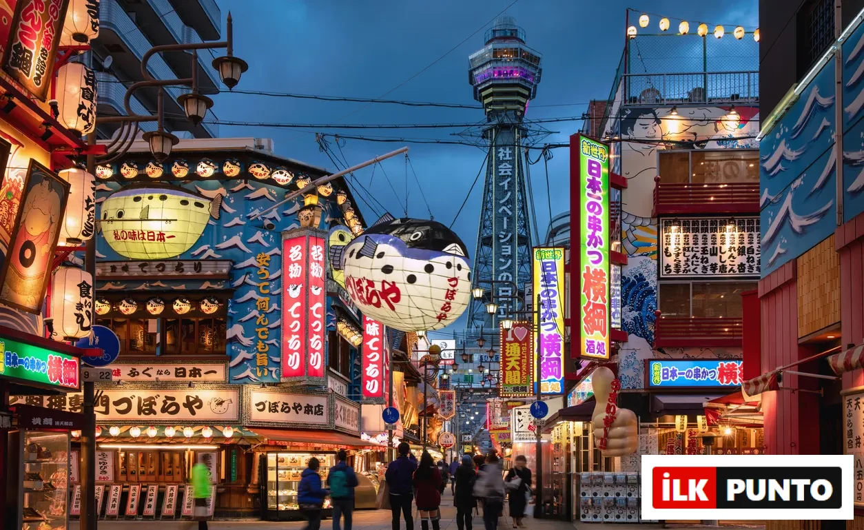 Osaka'nın Shinsekai bölgesinde, akşam alacakaranlığında neon ışıklı reklam panoları, devasa kirpi balığı fenerleri ve arka plandaki ikonik Tsutenkaku Kulesi'nin aydınlattığı kalabalık bir sokakta insanlar yürüyor.