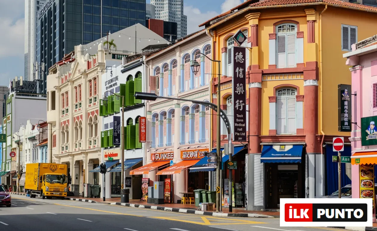 Singapur'un hareketli caddelerinden birinde, geleneksel mimariye sahip, kemerli pencereleri ve süslü cepheleriyle dikkat çeken sıralı, çok renkli dükkan binalarının bulunduğu, arka planda modern gökdelenlerin yükseldiği, tarih ve çağdaşlığın iç içe geçtiği özel bir şehir manzarası.