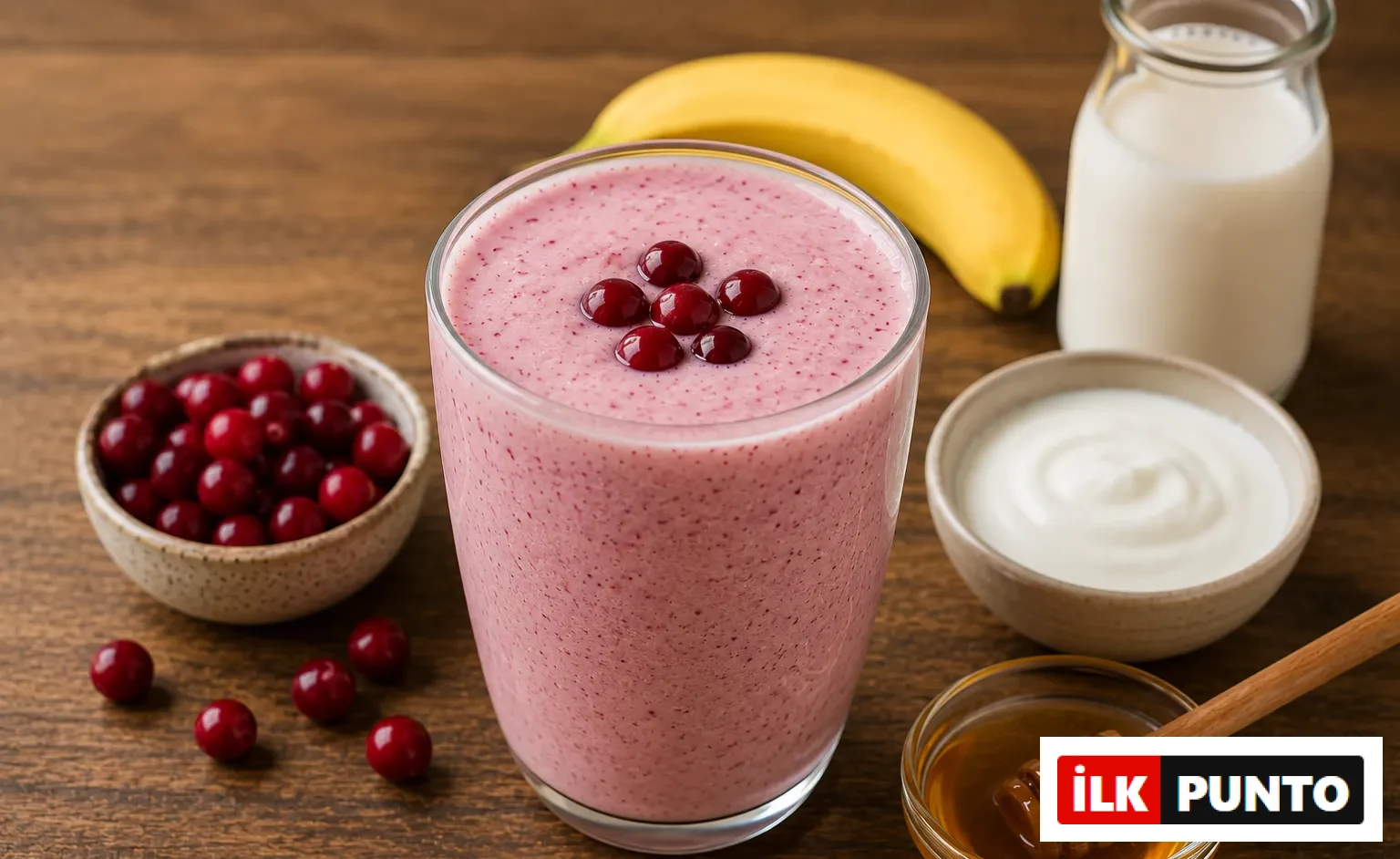 Ahşap bir masa üzerinde, altı parlak kırmızı kızılcıkla süslenmiş, pembe tonlarında pürüzsüz bir kızılcık smoothie bardağı, etrafında taze kızılcıklar, sarı bir muz, bir şişe süt, kremalı bir kase yoğurt ve ahşap bal kaşığıyla bal, sağlıklı ve lezzetli bir içecek sunumu.