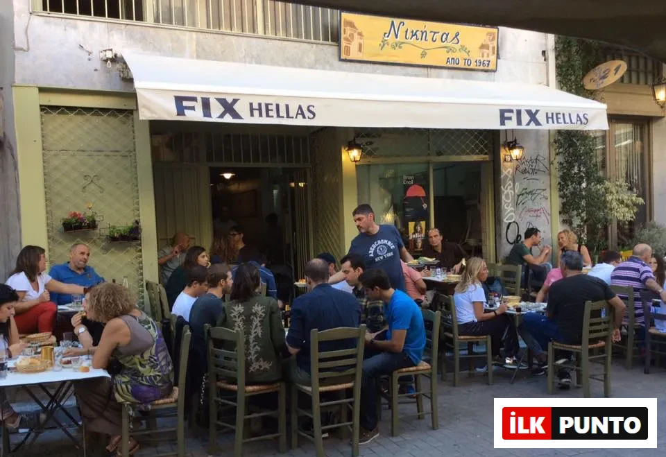 Beyaz bir tente üzerinde FIX HELLAS yazan, Nikitas adlı Yunan tavernasının önündeki kaldırımda, kalabalık bir grup insan yeşil sandalyelerde oturmuş, garsonun servis yaptığı masalarda sohbet edip yemek yiyerek keyifli bir açık hava mekanı atmosferi sunuyor.