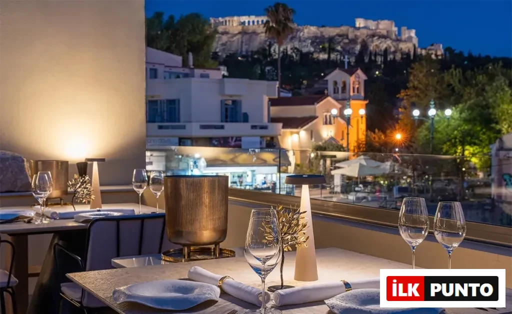 Atina'daki lüks bir restoranın terasında, masalar şık bir şekilde hazırlanmış; beyaz tabaklar, kadehler ve modern masa lambalarıyla donatılmışken, arka planda Akropolis'in gece ışıklarında büyüleyici silueti ve şehrin ışıltılı binaları görülüyor.