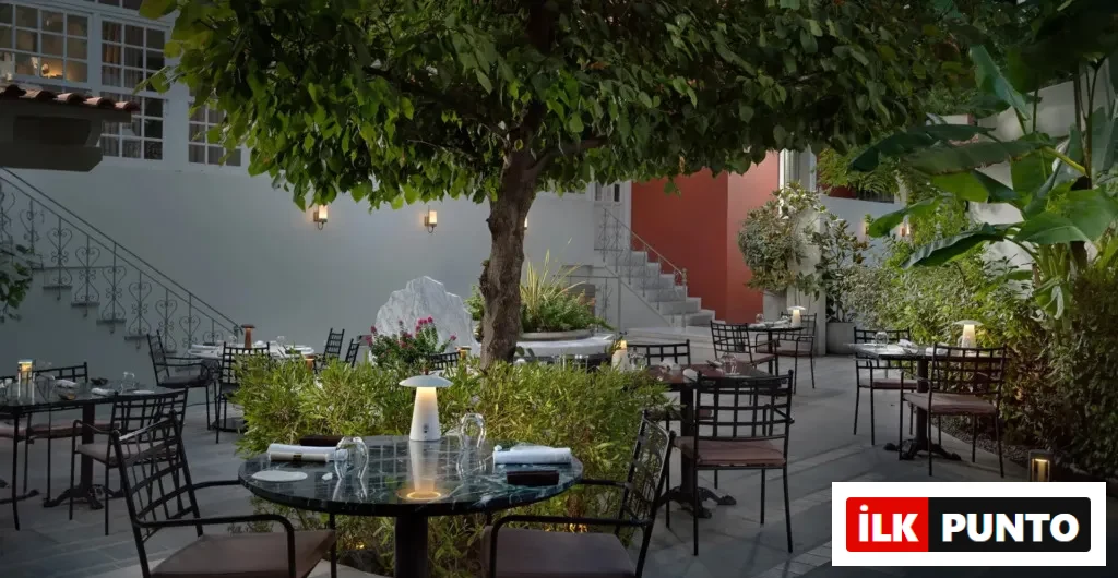 Bu resimde, geniş yapraklı bir ağacın altında, etrafı yeşil bitkilerle çevrili, mermer masa ve metal sandalyelerle düzenlenmiş şık bir açık hava restoranı alanı gösteriliyor, arka planda duvar lambalarıyla aydınlatılmış beyaz bir bina ve demir parmaklıklı merdivenler genel atmosfere huzurlu bir hava katıyor.