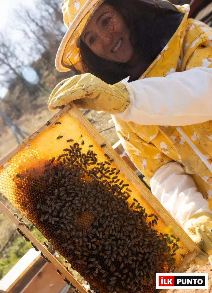 Sarı arıcı kıyafeti ve yüzünü koruyan tülüyle gülümseyen bir kadın, birçok arının çalıştığı ve parlak altın sarısı bal ile dolmaya başlayan petekli ahşap bir çerçeveyi özenle tutarak bal üretimi hakkında bilgi veriyor gibi görünüyor.