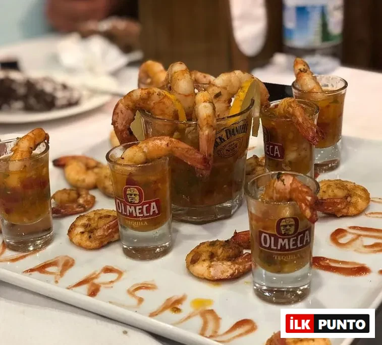 Beyaz dikdörtgen bir servis tabağında, içlerinde Jack Daniel's ve Olmeca Tequila marka shot bardakları da bulunan bardaklara ve tabağa özenle yerleştirilmiş baharatlı pişmiş karidesler, tabağın üzerinde sosla yazılmış ARSAN kelimesiyle Foça'daki bir restoranın dikkat çekici deniz ürünleri sunumunu yansıtıyor.