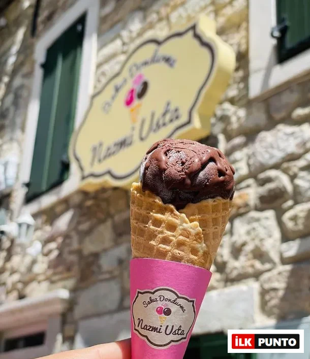 Pembe ambalajlı waffle külahında, hafifçe eriyerek aşağı doğru akan tek top koyu renk çikolatalı dondurmayı tutan ojeli bir el görünüyor; arka planda bulanık şekilde bir taş bina ve Sakız Dondurma Nazmi Usta yazılı sarı dükkan tabelası seçilirken, bu kare Nazmi Usta'nın meşhur dondurmalarından birini tadarken yaz gününün keyfini çıkaran bir anı yakalıyor.