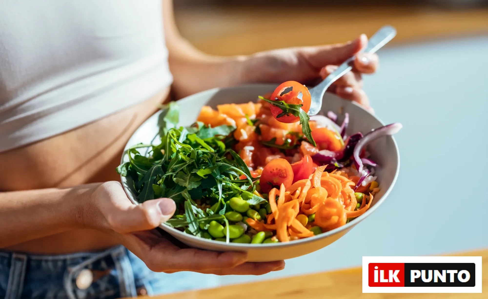 Beyaz bir atlet ve kot pantolon giyen bir kişi, taze roka, kiraz domates, rendelenmiş havuç, edamame ve kırmızı soğan içeren rengarenk, sağlıklı bir salata kasesini tutuyor, çatalında bir parça domates ve roka var, bu görüntü Başak burcu için önerilen Nisan ayı detoks enerjisi ve sağlık temasına uygun beslenmeyi vurguluyor.