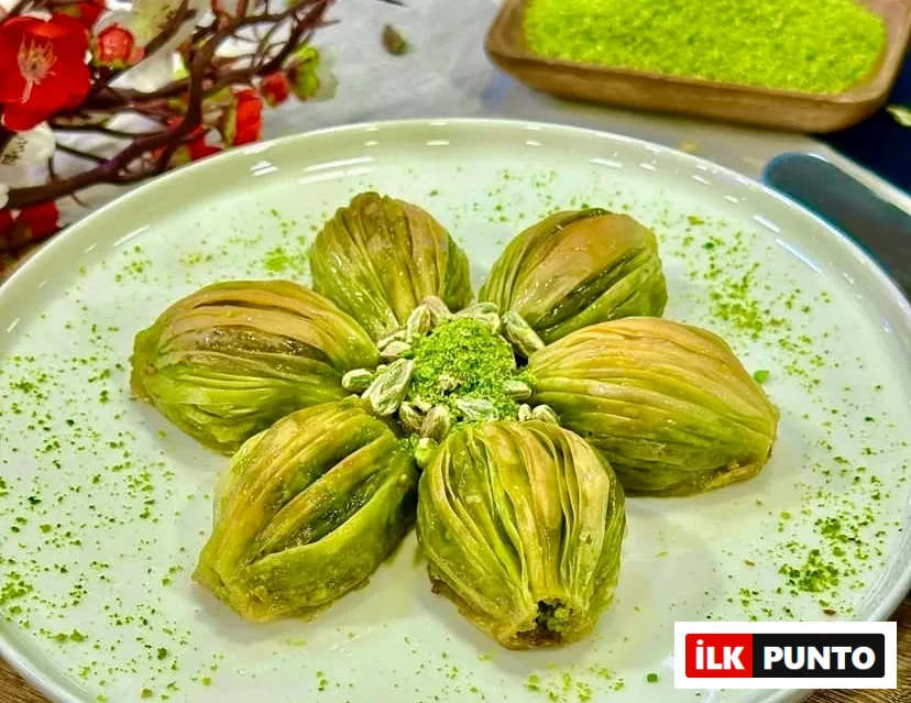 Beyaz tabakta çiçek formunda dizilmiş altı adet bol fıstıklı midye baklava. Merkezde Antep fıstığı tozu ve taneleri yer alırken, tabağın çevresine yeşil fıstık serpiştirilmiş. Arka planda kırmızı çiçekler ve fıstık dolu ahşap kaseyle