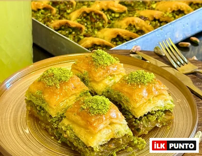 Altın sarısı tabakta sunulan, üzeri ve içi bol yeşil fıstık dolu dört dilim çıtır şerbetli baklava. Ahşap zeminli sunumun arka planında bir tepsi fıstıklı midye baklava, serinletici bir içecek ve şık çatal bıçak takımı yer