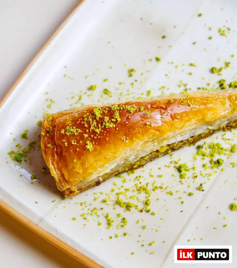 Beyaz seramik tabakta sunulan, ince açılmış yufkasıyla altın sarısı çıtır havuç dilimi baklava. Tatlının arasındaki kalın Antep fıstığı dolgusu öne çıkarken, parlak yüzeyine ve tabağın geneline ince çekilmiş yeşil fıstık tozu