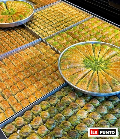İlk Punto Türkiye'nin En İyi Baklavacıları rehberindeki geleneksel lezzetleri yansıtan tepsiler: Altın sarısı çıtır yufkalı klasik kare baklavalar, yuvarlak tepside bol fıstıklı üçgen havuç dilimi ve alt sırada içi fıstık dolgulu yeşil-sarı renkli midye baklavalar yan yana