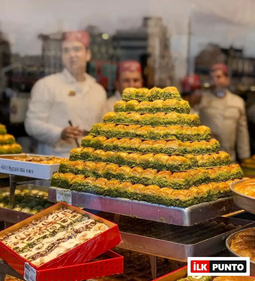 Türkiye’nin: İlk Punto Türkiye'nin En İyi Baklavacıları: Vitrinde piramit formunda dizilmiş bol fıstıklı çıtır baklava tepsisi sergileniyor. Alt kısımdaki kırmızı kutularda lokum çeşitleri yer alırken, bulanık arka planda beyaz üniformalı, kırmızı şapkalı ustalar geleneksel tatlıları hazırlıyor.