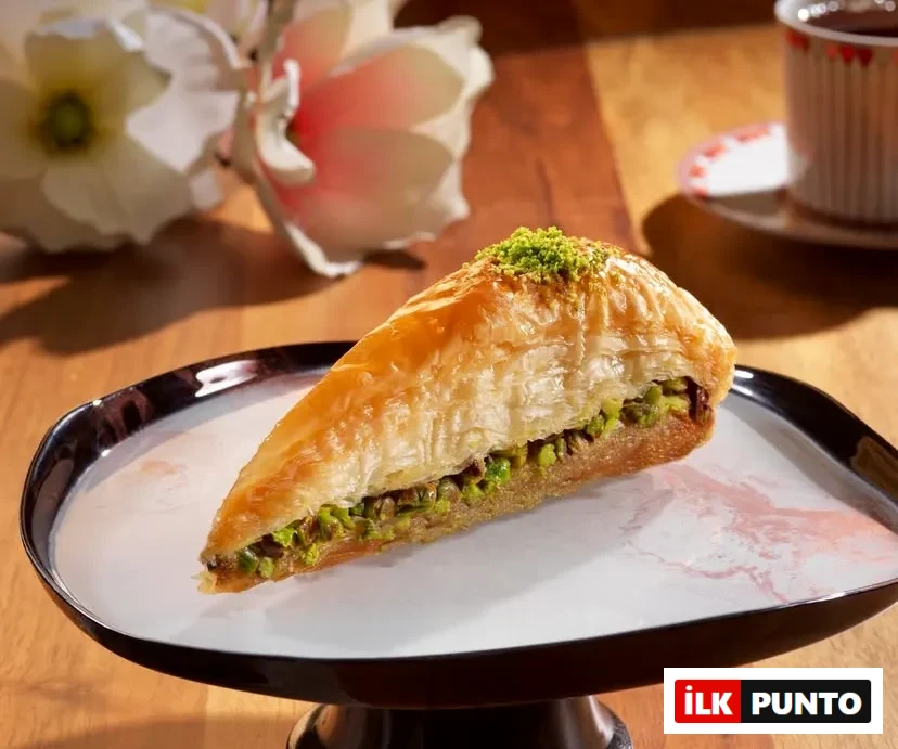 Türkiye'nin En İyi Baklavacıları rehberinde yer alan, incecik yufkalarla hazırlanan fıstıklı havuç dilimi baklava. Siyah ayaklı zarif beyaz bir tabakta sunulan bu çıtır geleneksel lezzete, ahşap masa üzerinde beyaz çiçekler ve ince belli bardakta taze demlenmiş Türk