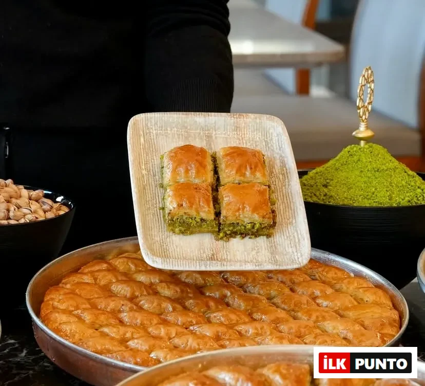 Siyah giyimli biri kare tabakta dört dilim fıstıklı çıtır baklava sunuyor. Alt kısımda metal tepsilerde nar gibi kızarmış şerbetli baklavalar, yanlarda ise taze bütün ve ince çekilmiş toz Antep fıstığı dolu siyah kaseler sergileniyor.