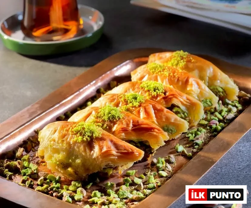 Bakır tepside sıralanmış, bol fıstık dolgulu ve üzeri Antep fıstığı tozuyla süslenmiş çıtır şöbiyet baklava dilimleri. Tepsi etrafına serpiştirilmiş fıstıklar ve arka planda ince belli bardakta geleneksel Türk çayı