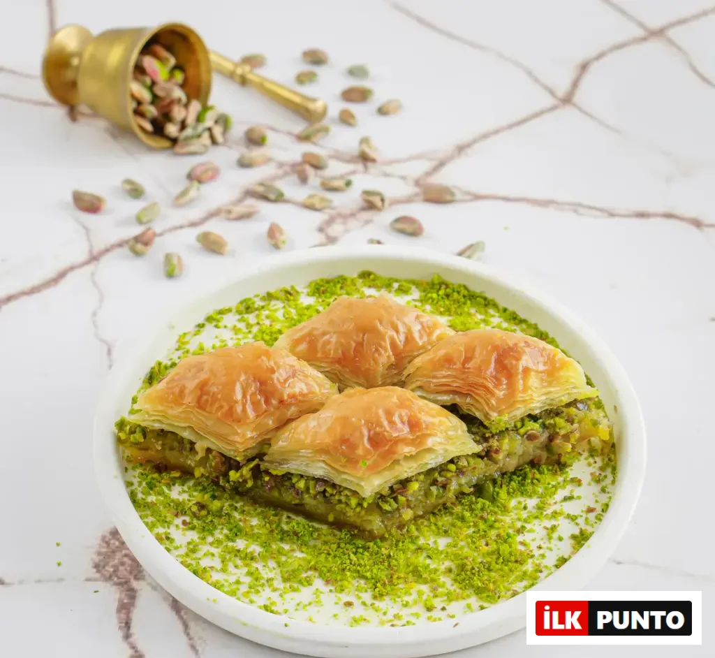 Beyaz mermer zemin üzerinde sunulan klasik fıstıklı baklava. Bolca serpilmiş yeşil toz fıstıkla kaplı beyaz tabakta dört dilim altın sarısı çıtır baklava yer alıyor. Arka planda devrilmiş pirinç bir havandan bütün Antep fıstıkları etrafa