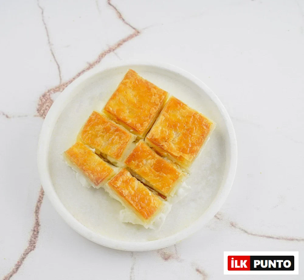 Beyaz mermer zemin üzerindeki tabakta sunulan, altın sarısı çıtır yufka katmanları ve beyaz kaymak dolgusuyla öne çıkan dilimlenmiş geleneksel tatlı. Bu enfes sunum, asırlık Türk tatlı kültürünün incelikli el işçiliğini ve zengin