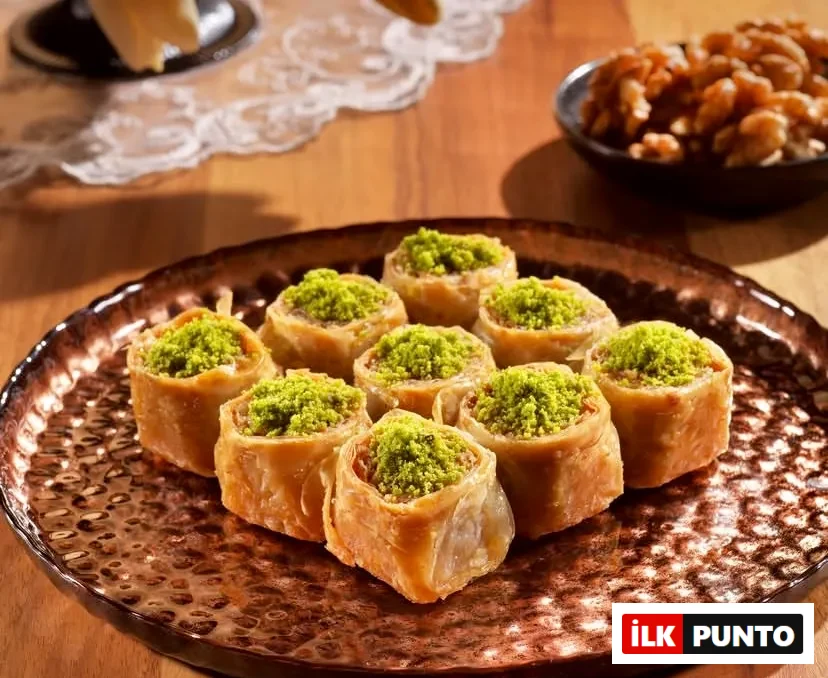 Dövme bakır tepside özenle dizilen, bol fıstıklı çıtır rulo baklavalar iştah açıcı biçimde sergileniyor. Arka plandaki ahşap masa, ceviz kasesi ve dantel detaylarıyla Türk mutfağının köklü geleneksel tatlı kültürü vurgulanıyor.