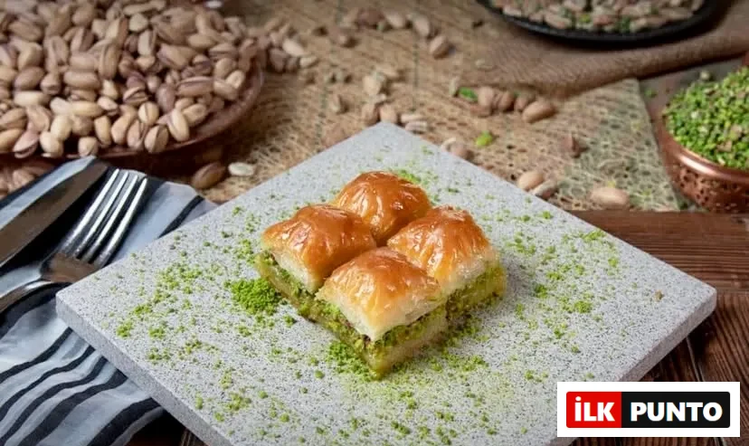 Türkiye’nin: İlk Punto, Türkiye'nin En İyi Baklavacıları: Ahşap masada gri taş tabakta sunulan, yeşil fıstık tozuyla süslenmiş dört dilim altın sarısı çıtır fıstıklı baklava. Arka plandaki taze Antep fıstıklarıyla zenginleşen bu sunum, Türk mutfağının ipek inceliğindeki köklü tatlı geleneğini
