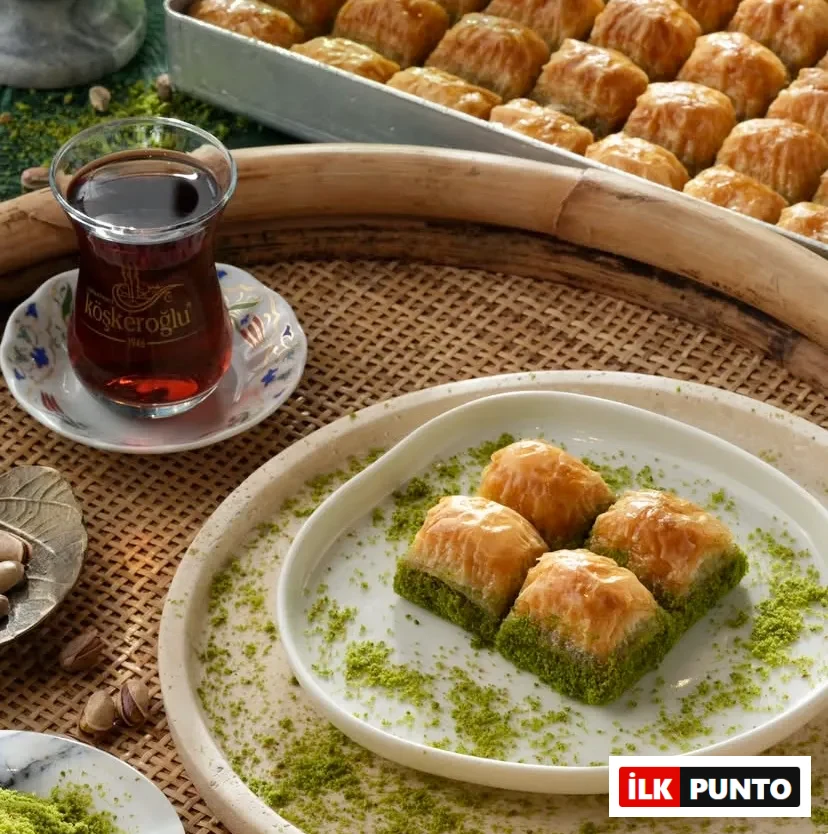 Köşkeroğlu logolu ince belli bardakta demli çay eşliğinde, beyaz tabakta sunulan dört dilim bol fıstıklı çıtır baklava. Hasır tepsiye serpiştirilmiş antep fıstığı tozları ve arka plandaki tepsi dolusu altın sarısı tatlılar iştah