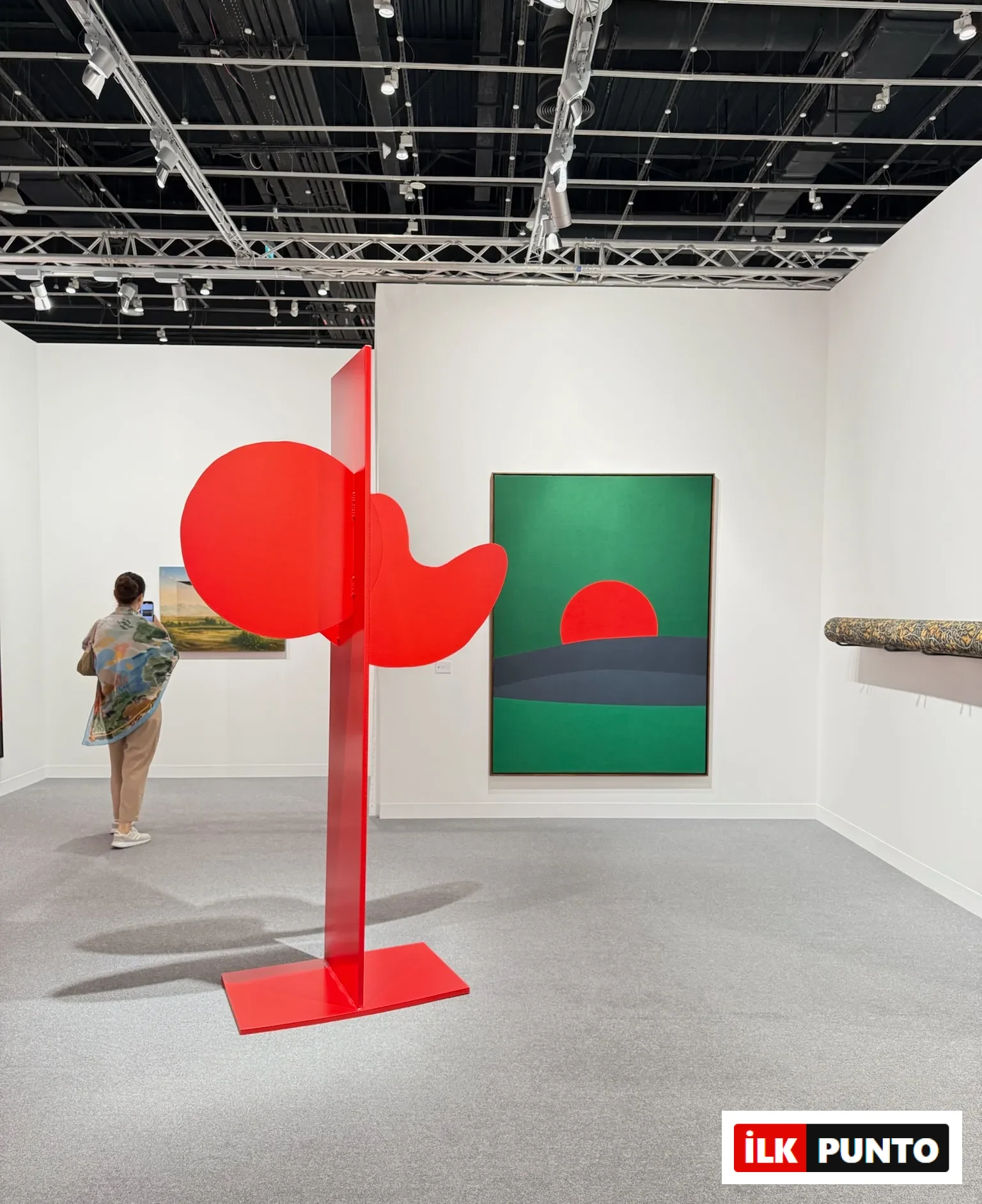 Abu Dhabi Art’ta kırmızı heykel yerleştirmesi ve arka planda geometrik kompozisyonlu çağdaş resimlerin yer aldığı sergi alanı