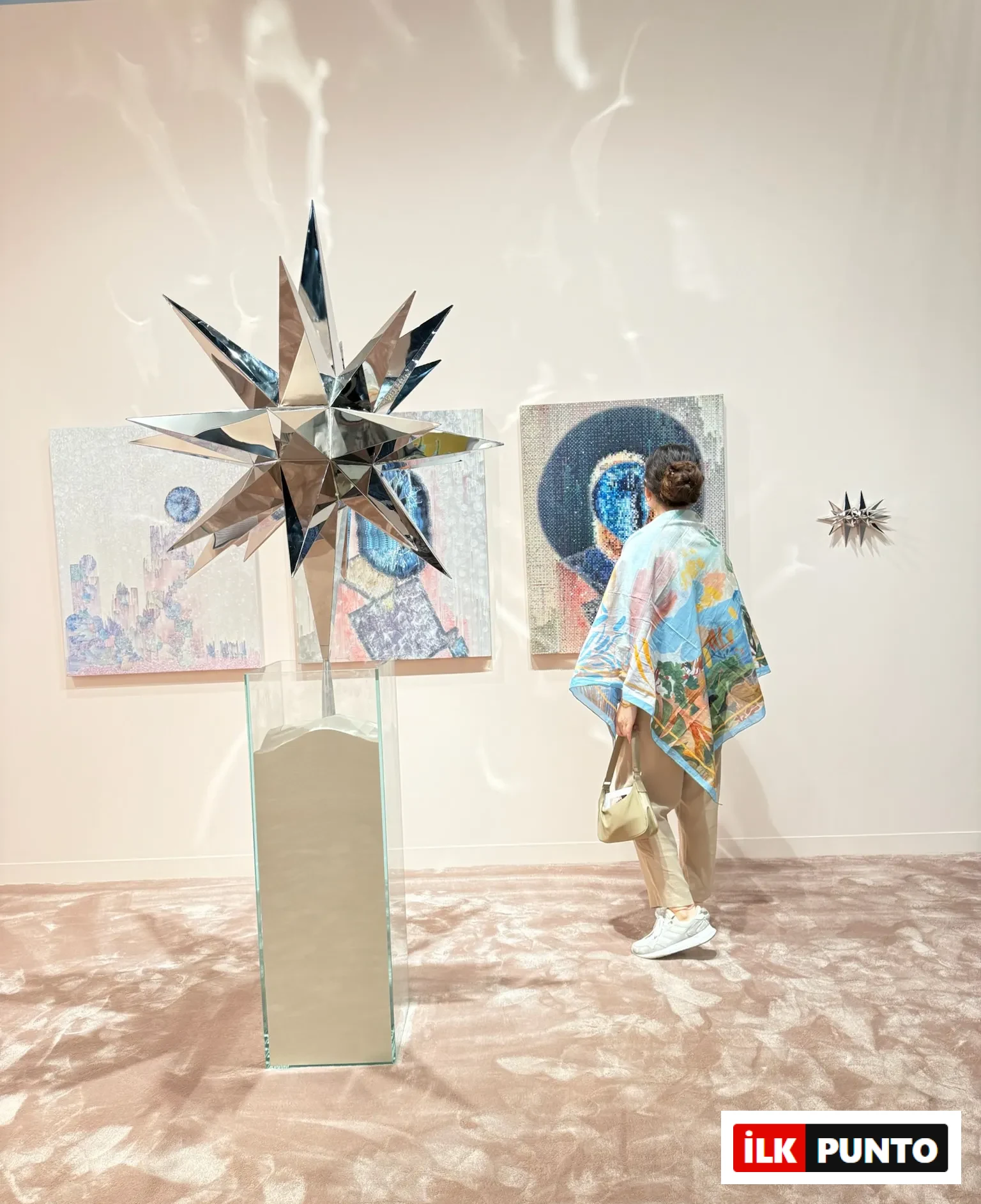 Abu Dhabi Art’ta ayna yüzeyli yıldız formunda bir heykel ve arka planda çağdaş resimlerin yer aldığı sergi alanını inceleyen ziyaretçi