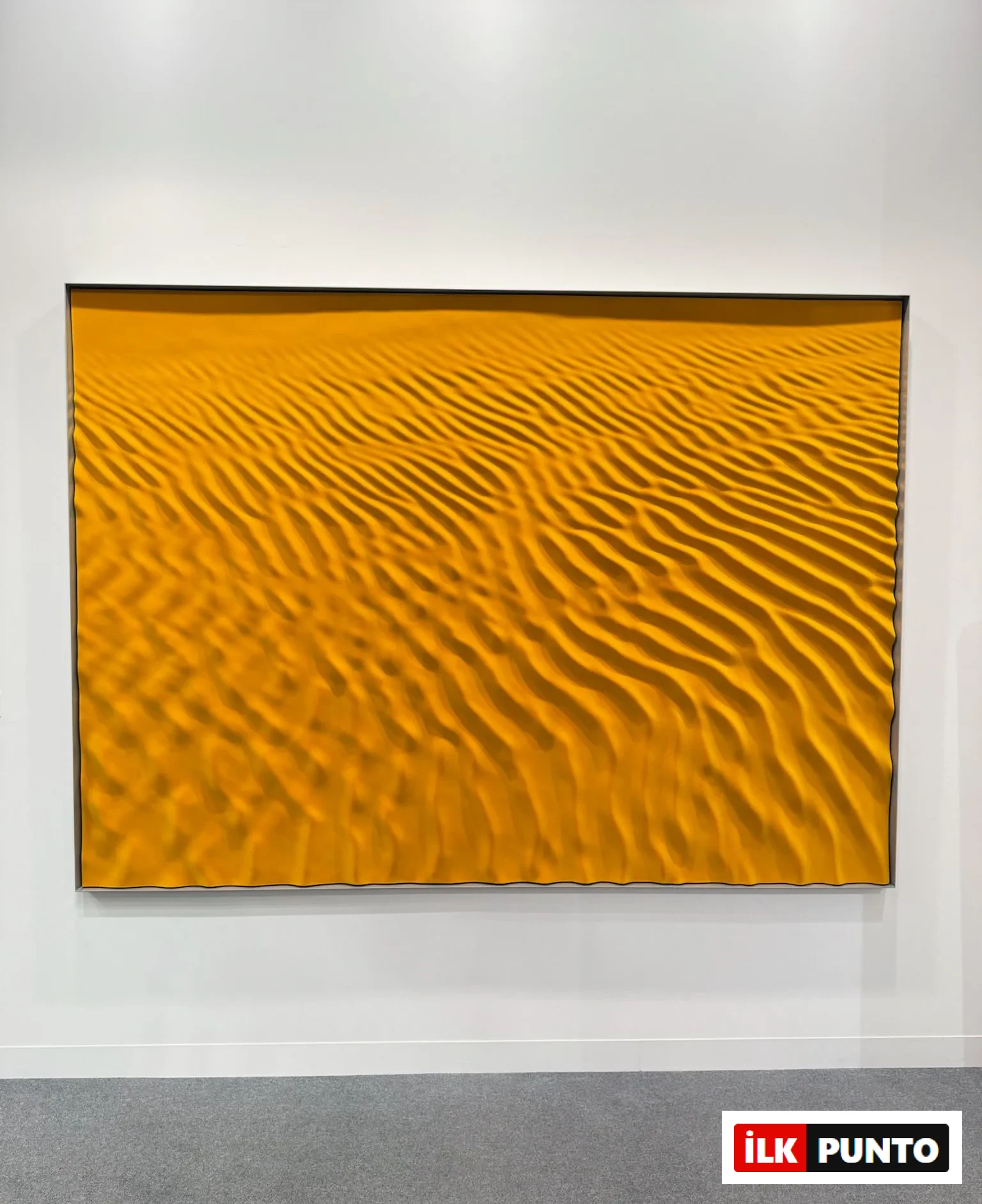 Abu Dhabi Art’ta sarı tonların hâkim olduğu, dalga dokulu soyut bir resmin sergilendiği galeri duvarı
