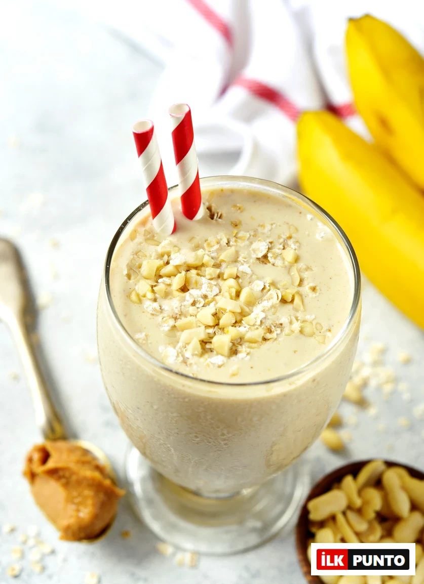 Bardakta, kırmızı beyaz çizgili iki pipetle servis edilen, soğuk buğulu camında parlayan, üzeri fıstık ve yulaf parçalarıyla süslenmiş Muzlu Fıstık Ezmeli Smoothie, mutfakta zaman kazandıran pratik ve lezzetli tarifler içeriğinin bir parçası olarak, yanında fıstık ezmesi ve bütün muzlarla iştah açıcı görünüyor.