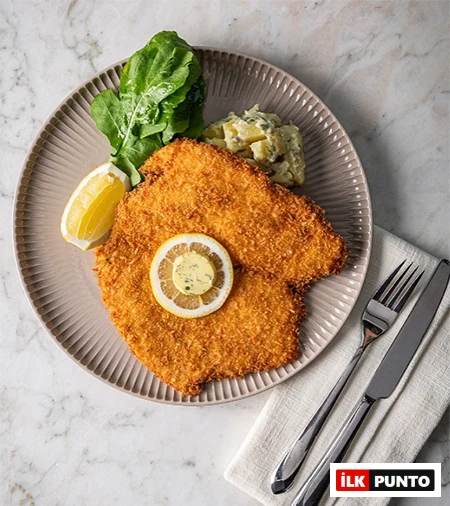 Bebek’in en gözde mekanları arasında konumlanan Divan Restaurant'tan, mermer zemin üzerinde zarif bir tabakta sunulan, tereyağlı limon dilimi, roka ve patates salatası eşliğindeki çıtır pane şnitzel yemeği, çatal bıçak takımıyla birlikte iştah açıcı bir görünüm sergiliyor.