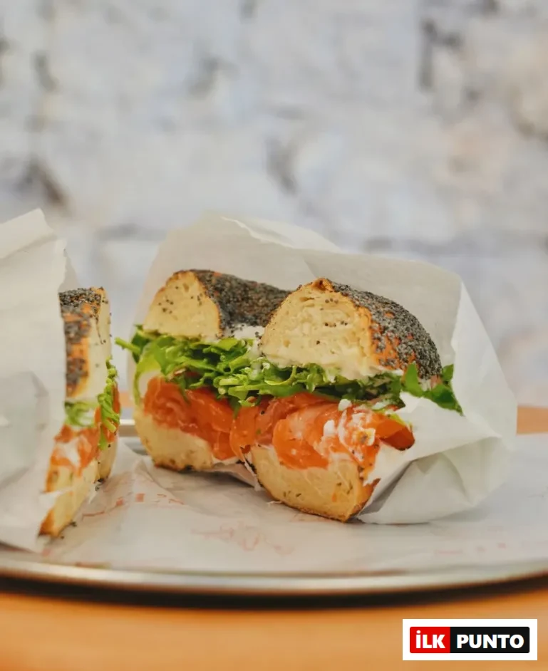 Gümüş bir tepside, beyaz ambalaj kağıdına sarılı, üzerine haşhaş tohumlarıyla süslenmiş, ortadan ikiye kesilmiş bir bagel sandviçinin içinden bol miktarda narince dilimlenmiş füme somon, krem peynir ve canlı yeşil roka yaprakları taşıyor; bu keyifli ve iştah açıcı lezzet, arka plandaki bulanık beyaz tuğla duvar önünde sunuluyor.