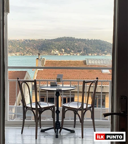 Bebek'teki en iyi mekanlardan birinin balkonundan, kırmızı kiremitli çatıların ve yemyeşil tepelerin arkasında uzanan masmavi Boğaz'ın büyüleyici manzarasına karşı yerleştirilmiş iki ahşap sandalyeli küçük bir masanın davetkar görüntüsü.