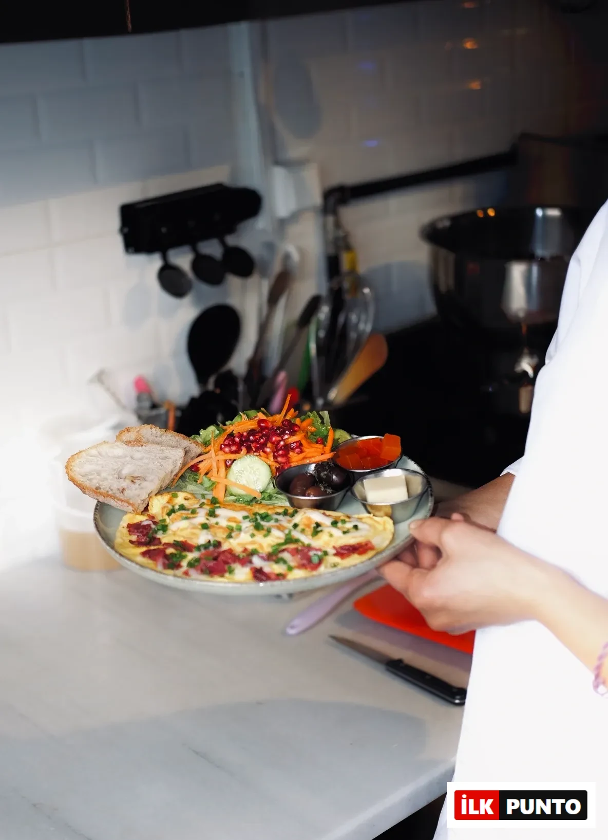 Ankara'daki Bala Bakehouse'tan beyaz önlüklü bir çalışan tarafından sunulan, sucuklu omlet, nar taneleriyle zenginleştirilmiş yeşil salata, kızarmış ekmek dilimleri, zeytin, reçel ve tereyağı içeren rengarenk ve doyurucu bir kahvaltı tabağı.