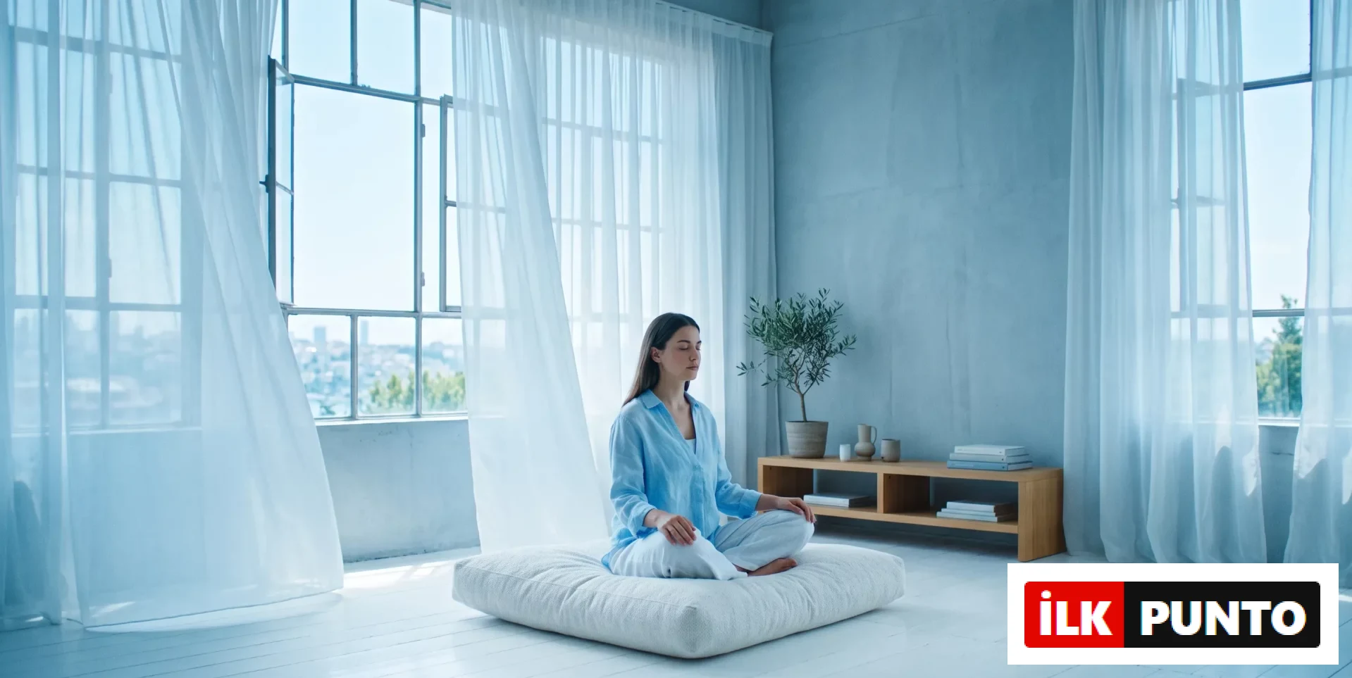 Ortada bağdaş kurmuş, gözleri kapalı, açık mavi gömlekli bir kadın, ferah ve aydınlık bir odada büyük pencerenin önündeki beyaz minder üzerinde meditasyon yaparak huzur bulmaya çalışıyor; hafifçe dalgalanan şeffaf perdeler ve odanın sakin atmosferi, stres yönetimi için ideal bir ortam sunuyor.