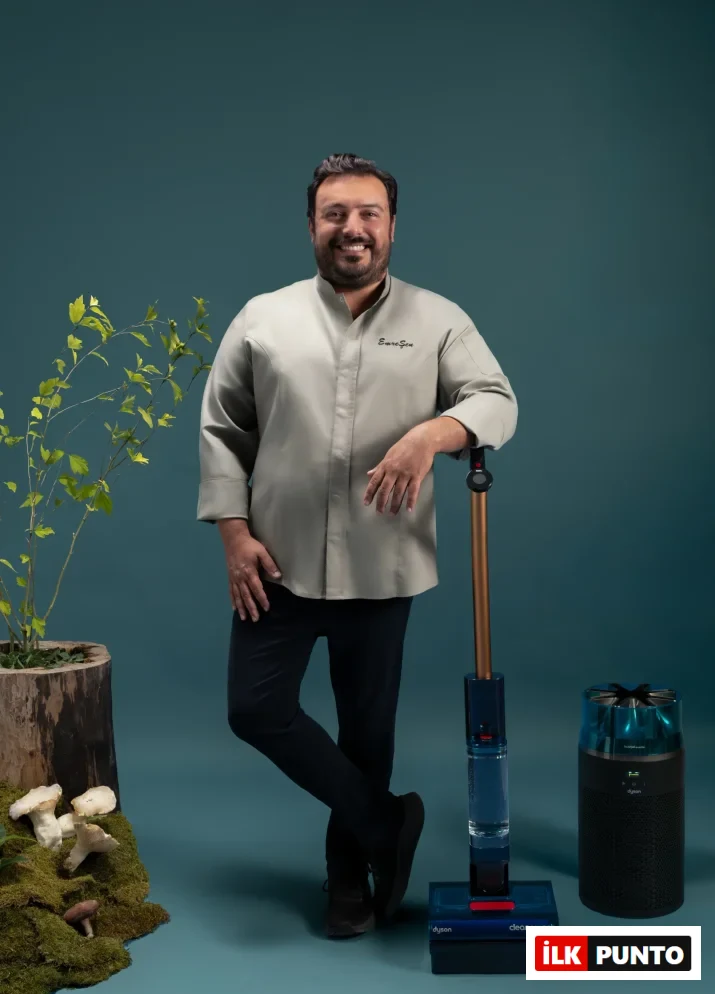Açık gri şef ceketi giyen, gülümseyen ve kameraya bakan Emre Şen, sol kolunu üzerinde Dyson Clean+Wash yazısı görünen ıslak ve kuru zemin temizleyicisine yaslamış; yanında siyah renkli bir Dyson hava temizleyici dururken, sol tarafında ahşap kütük saksıda yeşil yapraklı bir bitki ve yerden mantarlar yükselen doğal bir dekorun çevrelediği bu düzenlemede ev hijyeni ve temizlik teknolojilerini vurguluyor.
