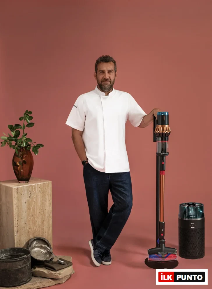 Beyaz şef ceketli ve kot pantolonlu Maksut Aşkar, gülümseyerek, sağ eliyle modern bir Dyson dikey süpürgeye yaslanmış; yanında bir Dyson hava temizleyici, arkasında ise geleneksel mutfak eşyaları ve bitkili bir vazo, hijyen ve çağdaş yaşam dengesini yansıtan bir kompozisyon oluşturuyor.