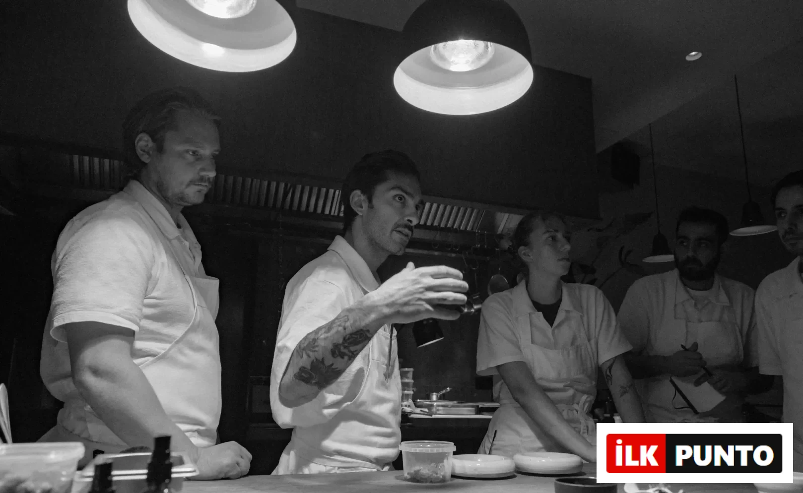 Amsterdam'daki bir restoran mutfağında, beyaz önlüklü şefler, ortadaki dövmeli şefin yemek hazırlığı veya yeni bir sunum tekniği üzerine elleriyle yaptığı detaylı açıklamayı ilgiyle dinlerken, tezgâh üzerinde yemek öğeleri ve servis kaseleri belirginleşiyor.