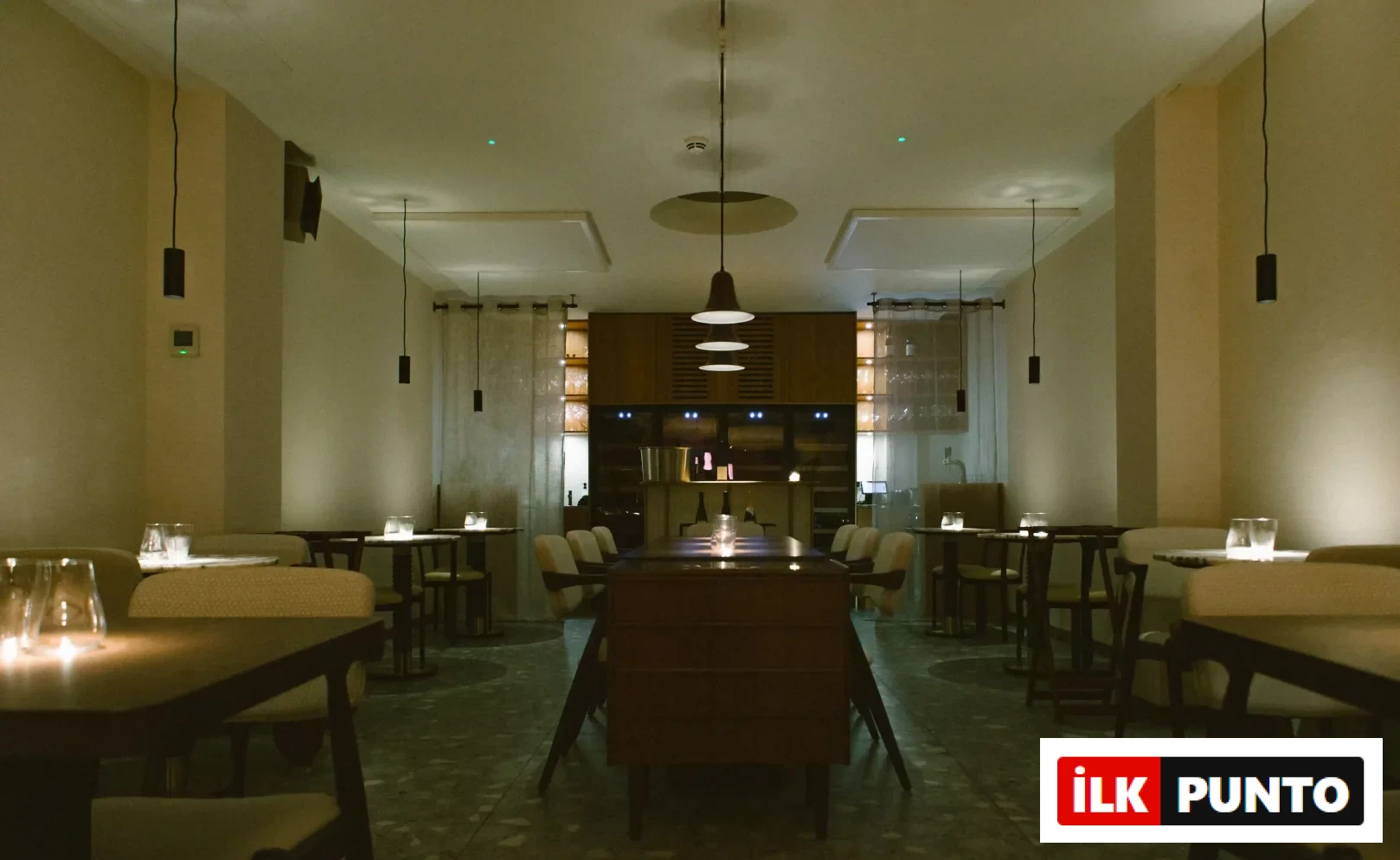 Loş ışıklı, modern tasarımlı bir restoranda, ortada uzun bir ahşap masa ve etrafında krem renkli sandalyelerle, duvar kenarlarında şık küçük masaların mum ışıklarıyla aydınlatıldığı, davetkar ve sakin bir yemek ortamı.