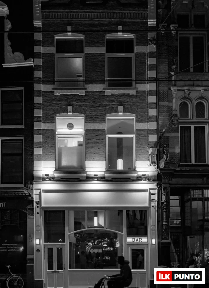 Amsterdam'da geceleri tuğla cepheli, ışıklarla aydınlatılmış bir barın önünden bisikletle geçen bir siluet, şehrin hareketli atmosferini ve gece hayatını yansıtan dinamik bir şehir sahnesi sunuyor.