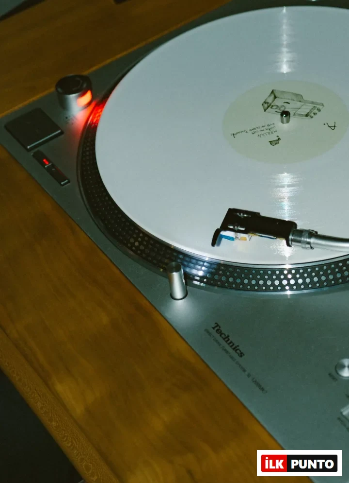 Üzerinde beyaz bir plağın bulunduğu, açma düğmesindeki kırmızı ışığıyla Technics SL-1200MK model bir pikabın ahşap bir yüzeyde konumlandırılmış hali yakın çekim olarak gösterilirken, plağın yüzeyine hassasça inmiş iğnesiyle detaylı bir dinleme deneyimine işaret ediyor.